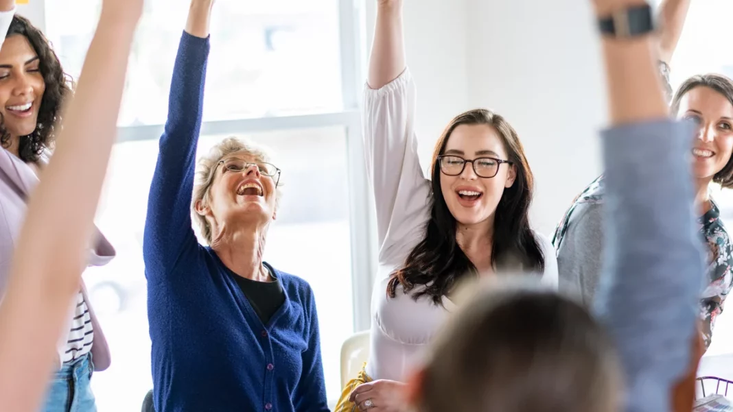 Women of different ages raising their hands in a meeting show why upskilling for women in leadership matter